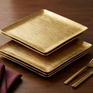 A stack of square gold textured plates neatly arranged on a long wood banquet table alongside a stainless steel buffet food warmer.