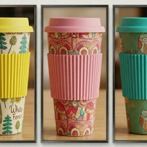 Set of three reusable bamboo fiber coffee cups featuring decorative printed designs, matching lids, and silicone grip sleeves in yellow, pink, and teal colors, displayed on a wooden surface.