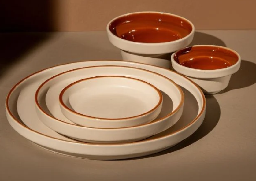 A close-up view of stacked off-white handcrafted ceramic plates and bowls with distinct natural terracotta rims, showing a hand with clear nail polish inspecting the texture.